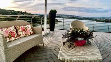 Ein Balkon mit Korbmöbeln, einem Blumentopf und Blick auf einen Fluss und Hügel.