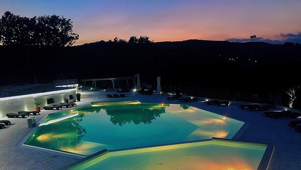A swimming pool with a geometric design, surrounded by lounge chairs and a building with a lighted roof.