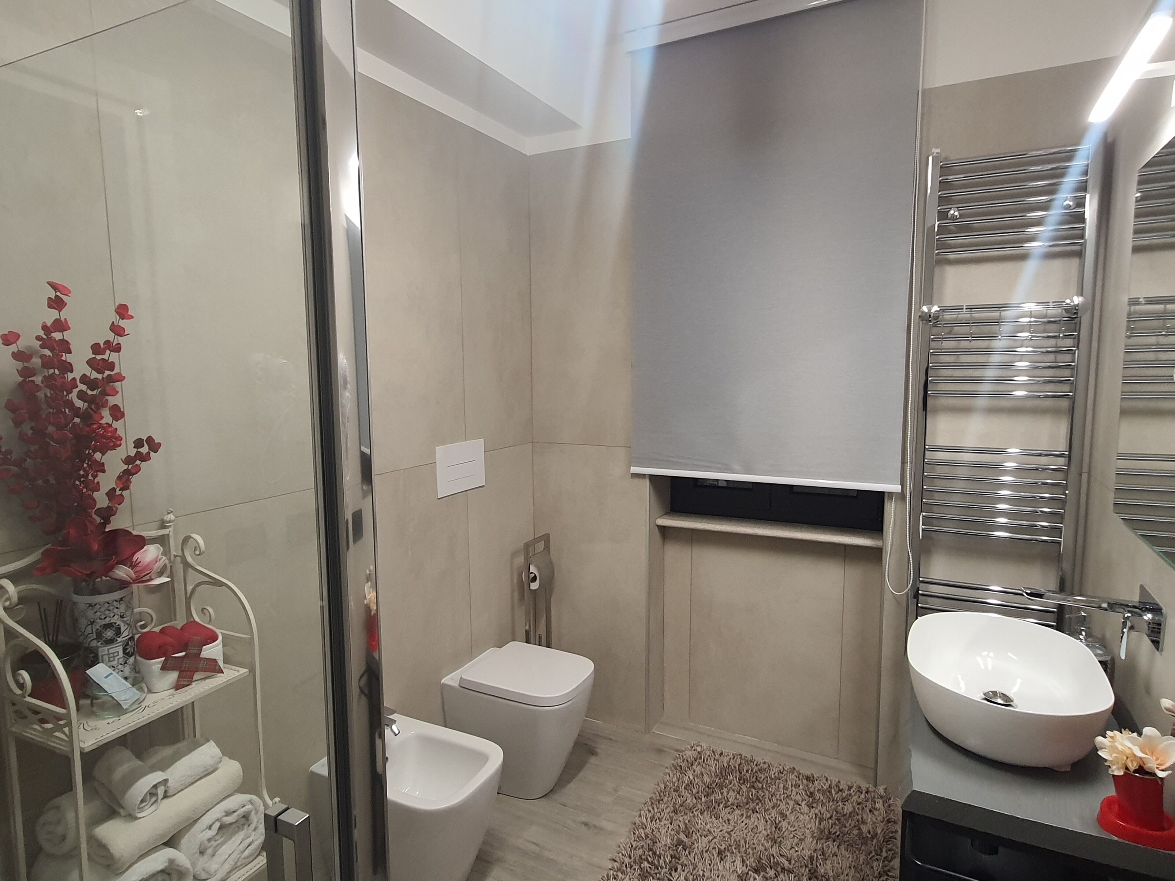 Un baño moderno con cabina de ducha de vidrio, inodoro blanco, bidé, lavabo blanco con recipiente y toallero con un florero rojo.