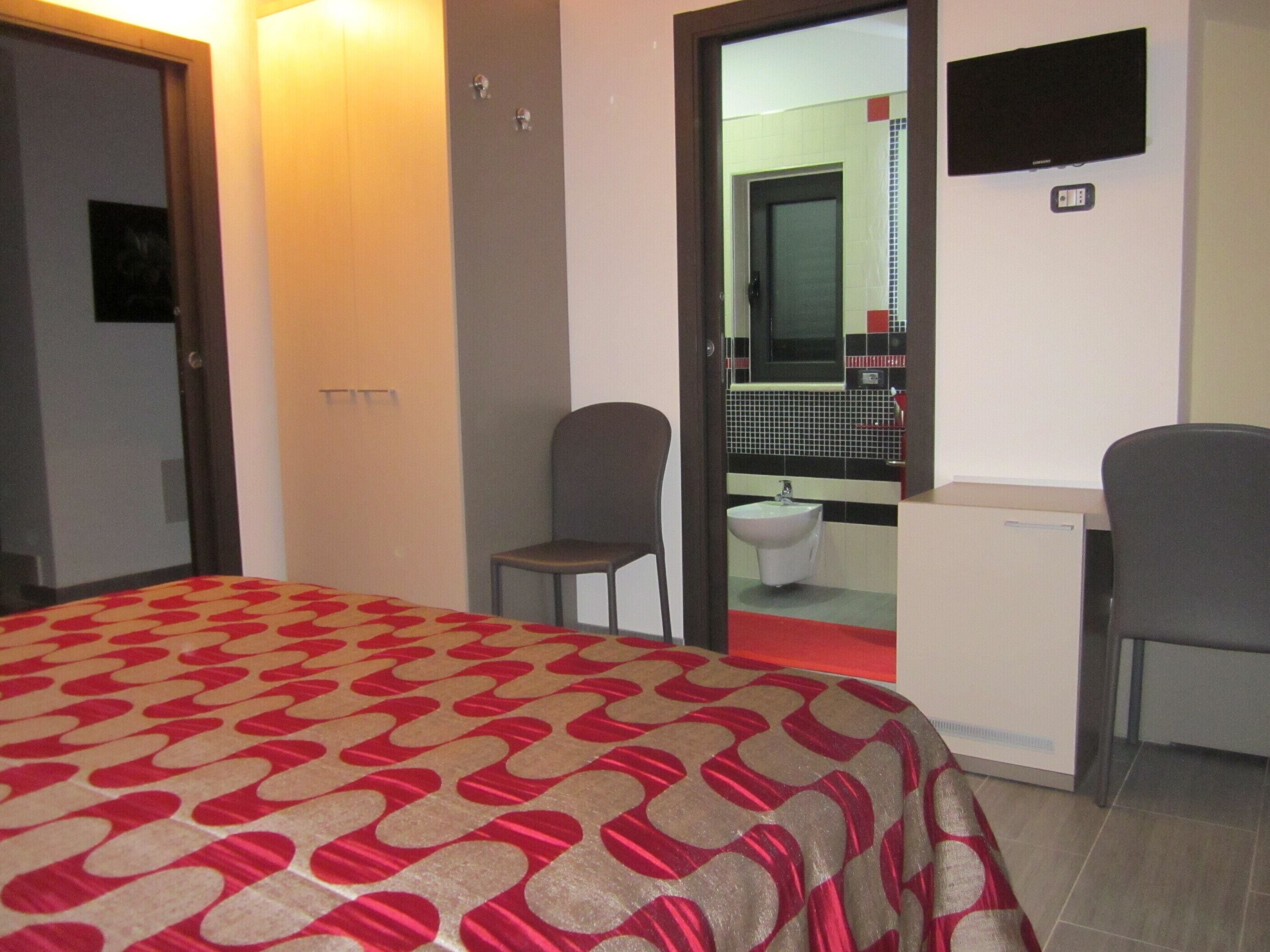 A hotel room with a bed, two chairs, a small desk, a television, and a bathroom visible through an open door.