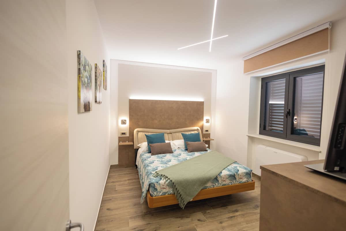Ein Schlafzimmer mit Bett, Nachttischen, einem Fenster mit Jalousien und einem Fernseher auf einem Schrank.
