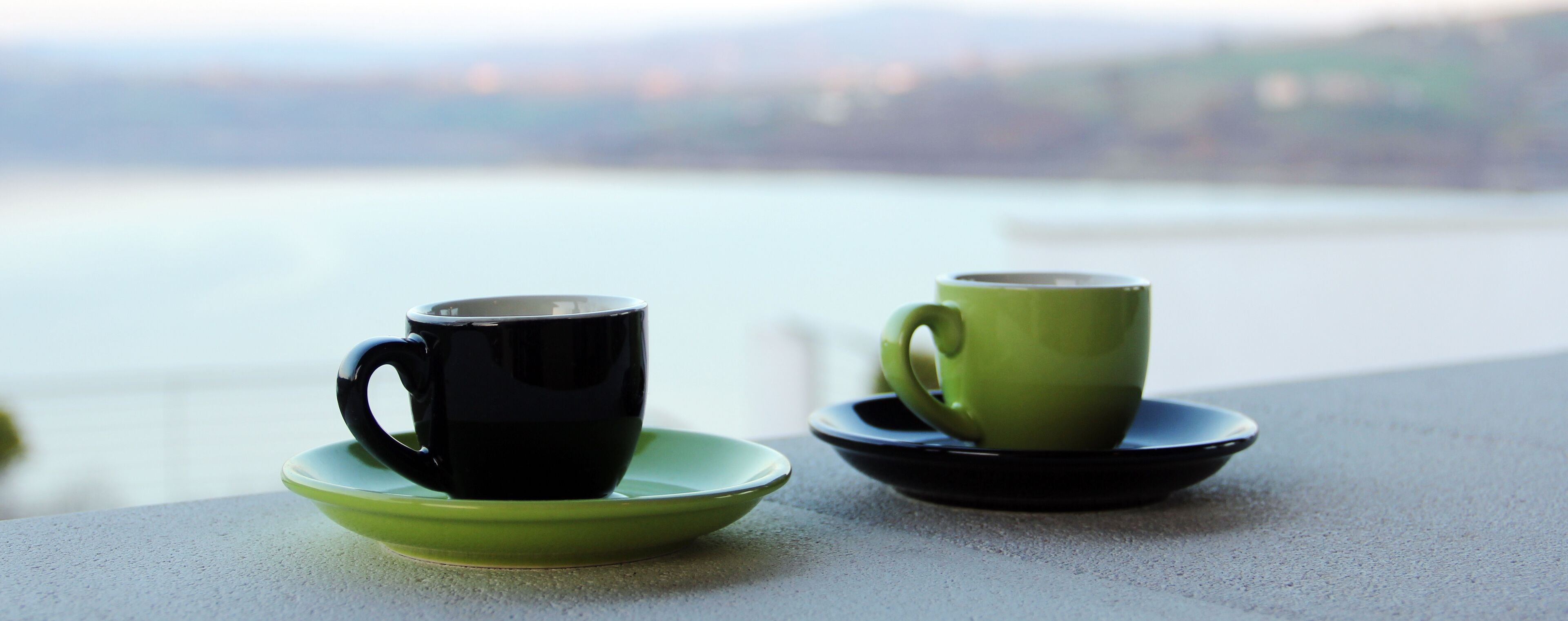 Twee kopjes op schoteltjes op een balkon met uitzicht op een wateroppervlak.