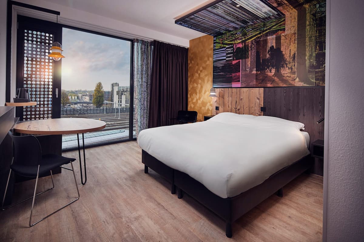 A modern hotel room with a large bed, a desk with a chair, a round table, and a large window offering a city view.