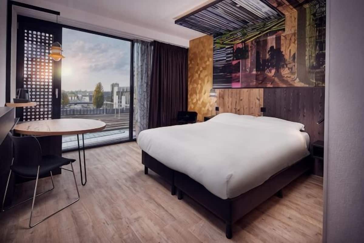 A modern hotel room with a large bed, a desk, a chair, a round table, and a city view through large windows.
