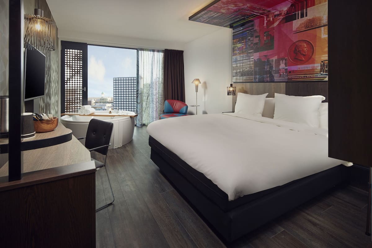A modern hotel room with a large bed, a desk with a chair, a small round table, and a large window offering a city view.