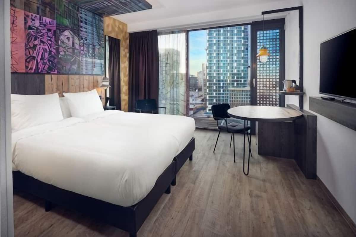 A hotel room with a large bed, a desk with a chair, a TV, and a view of a cityscape.
