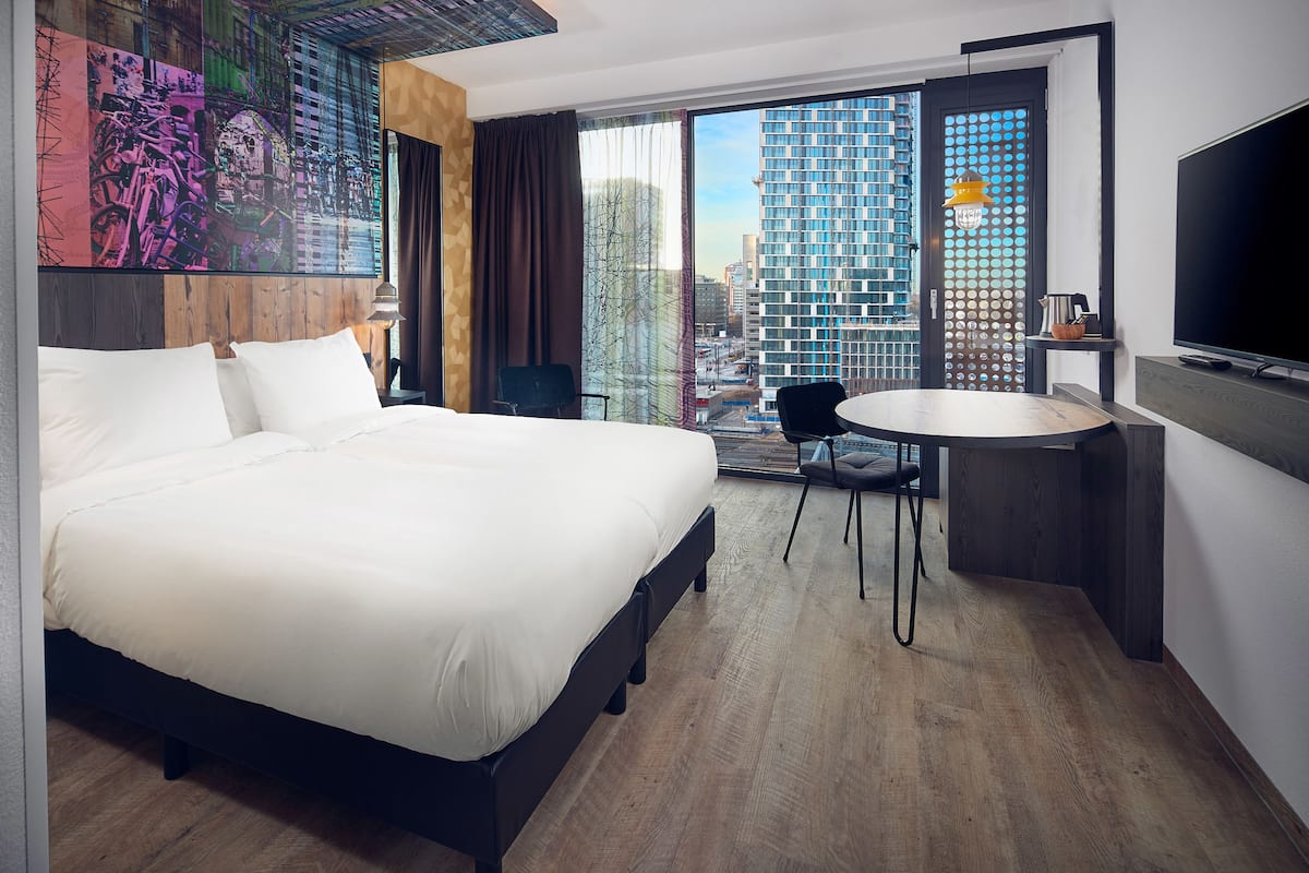 A hotel room with a large bed, a desk with a chair, a TV, and a city view through the window.