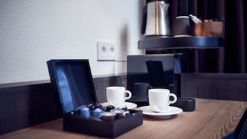 A hotel room with a wooden table, two white cups, a coffee maker, and a tray of chocolates.