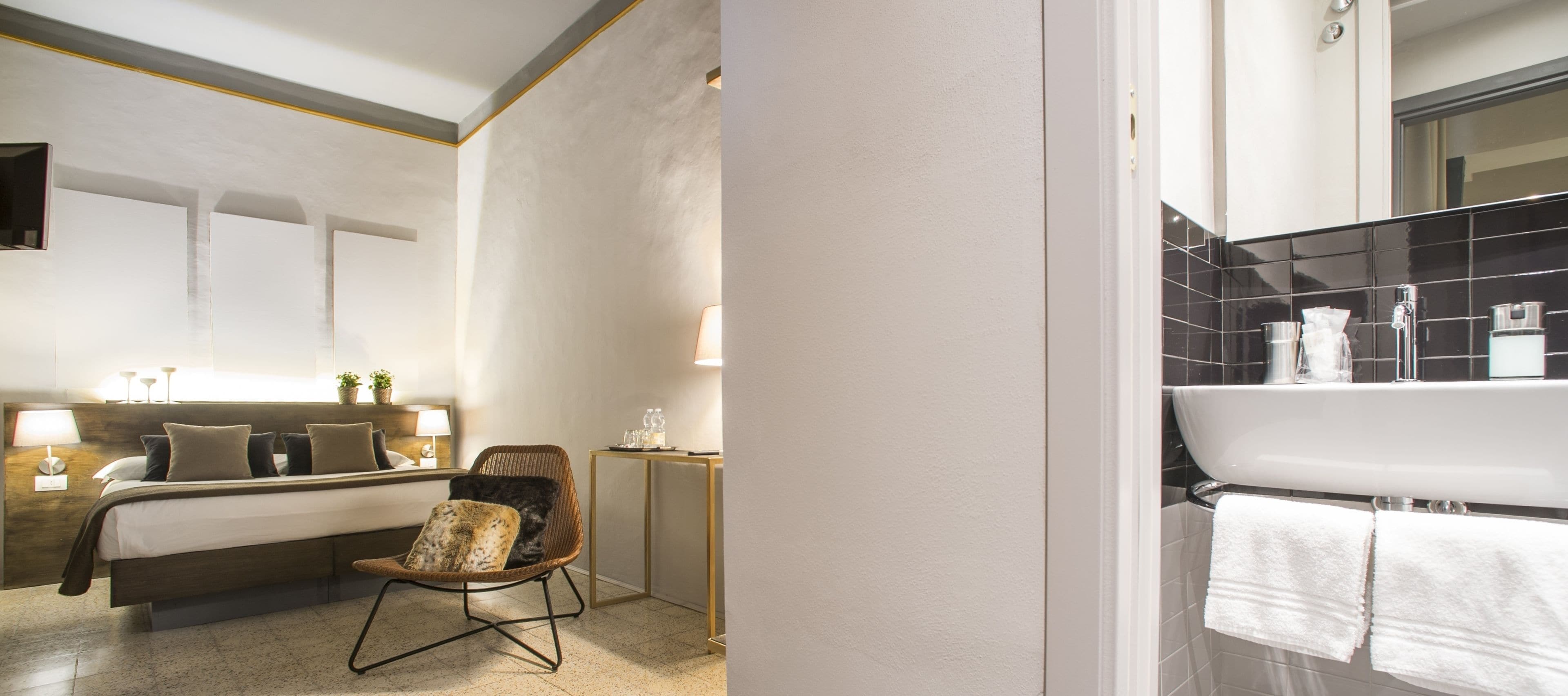 A hotel room with a bed, a chair, a bedside table with a lamp, a small table with toiletries, and a bathroom with a sink and towel rack.