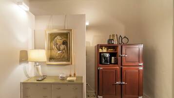 A hotel room with a bedside table, a lamp, a framed artwork, and a wooden cabinet with a coffee maker.