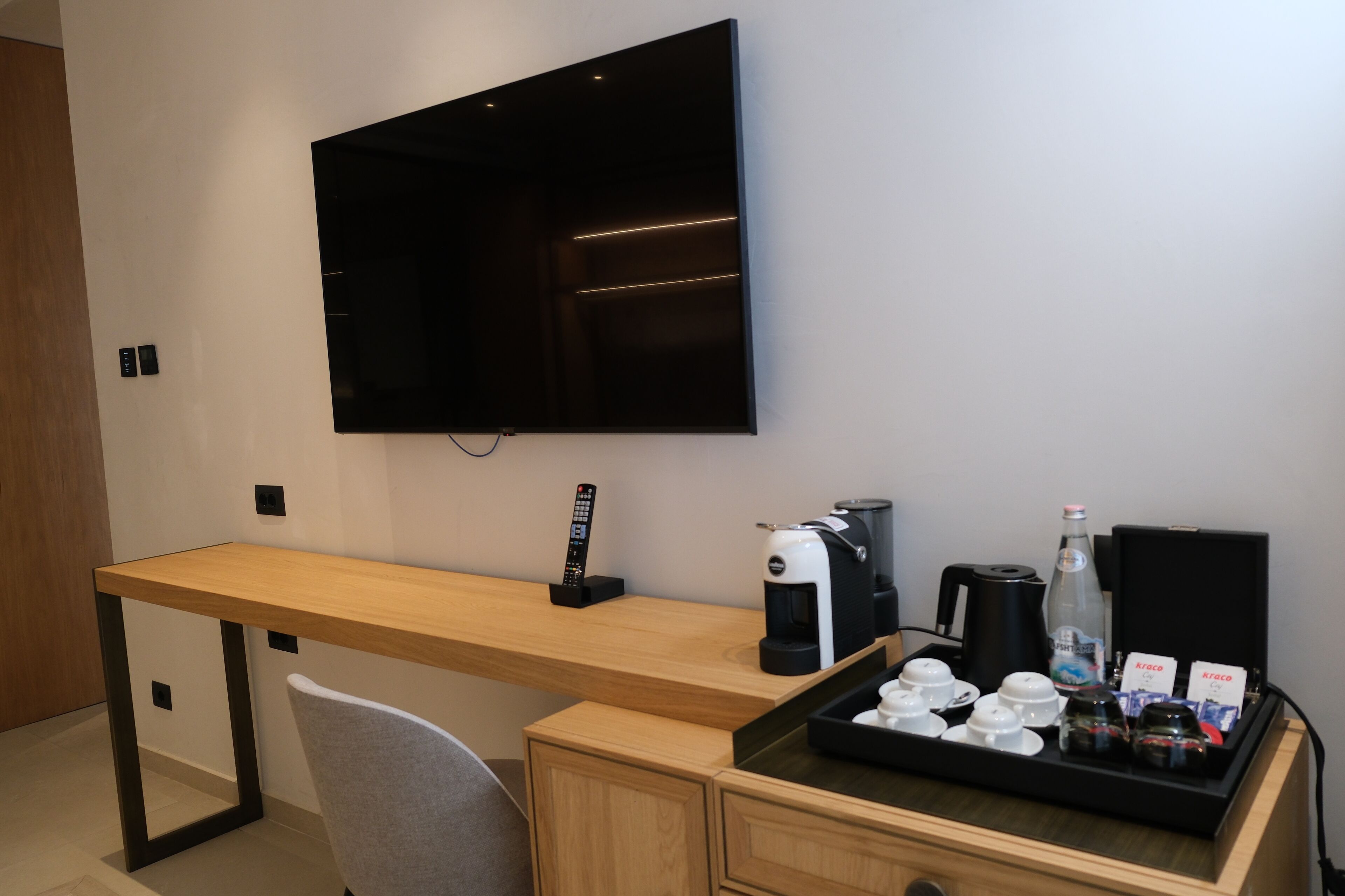 A modern hotel room with a wooden desk, a mounted flat-screen TV, a coffee maker, and a tray with cups and condiments.