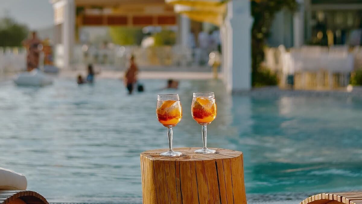 Two glasses of a beverage on a wooden stool by a poolside.