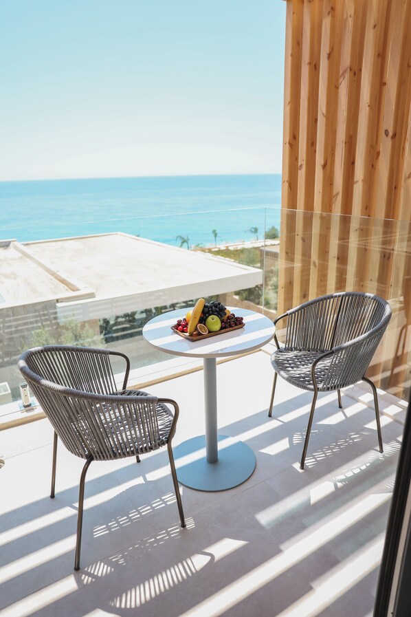 A balcony with two wicker chairs, a round table with fruit, and a view of the sea.