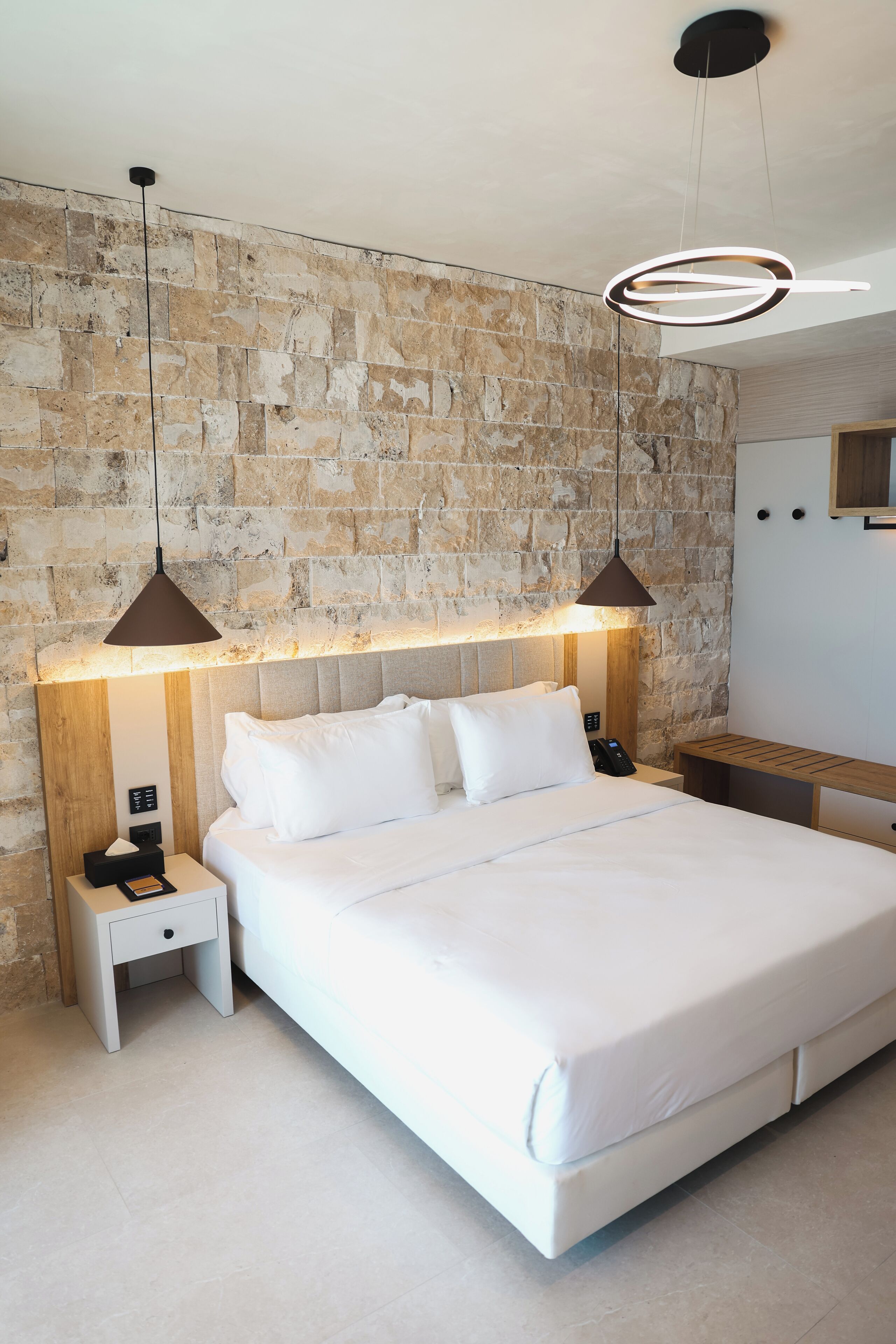 Habitación de hotel moderna con una pared de acento de piedra, una cama grande con ropa blanca, dos mesitas de noche e iluminación colgante.
