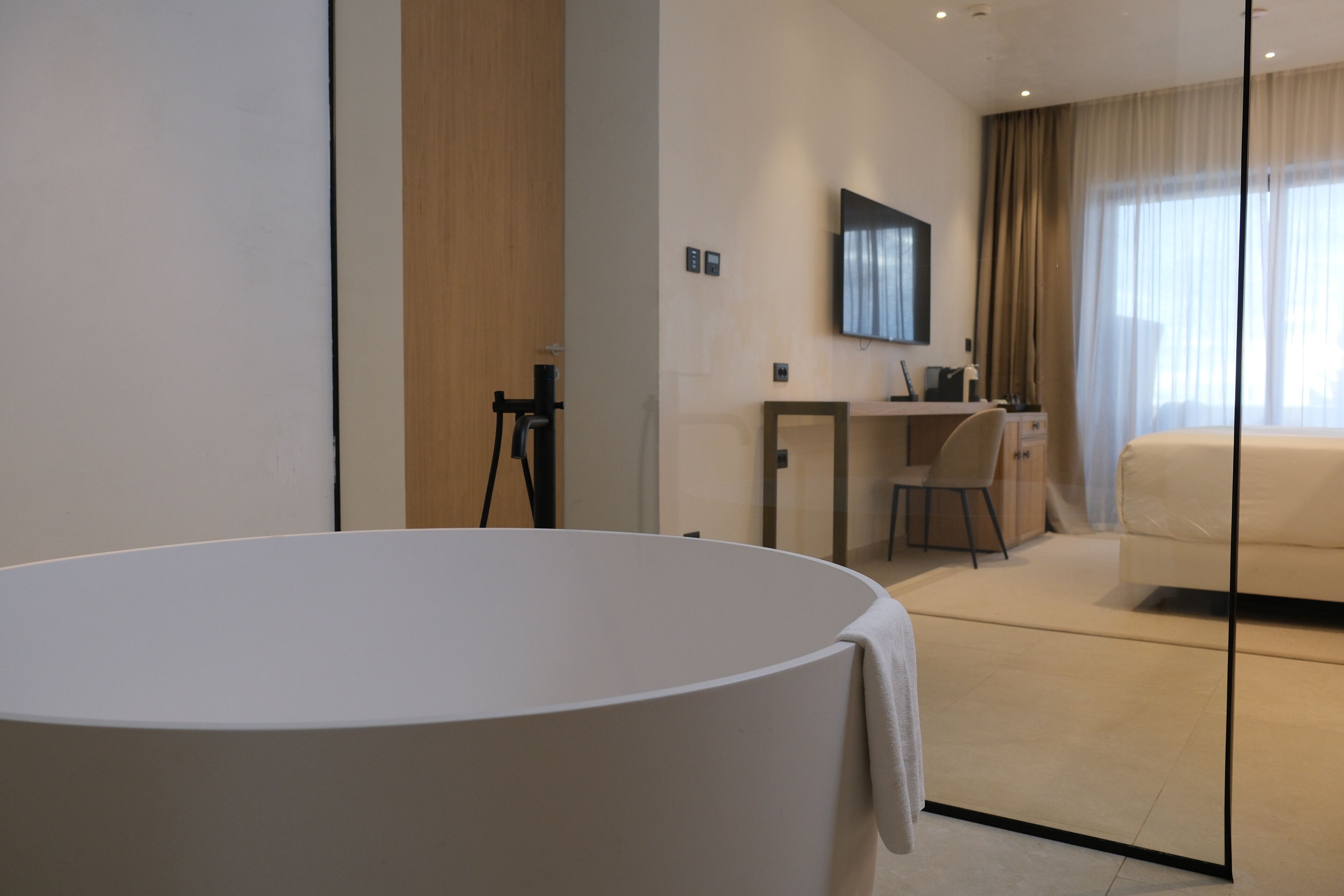 A modern hotel room with a large round bed, a desk with a chair, a television mounted on the wall, and a large window with curtains.