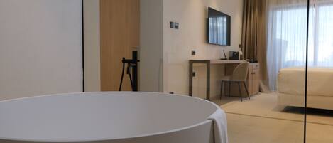 Een moderne hotelkamer met een groot rond bed, een bureau met stoel, een aan de muur bevestigde televisie en een groot raam met gordijnen.