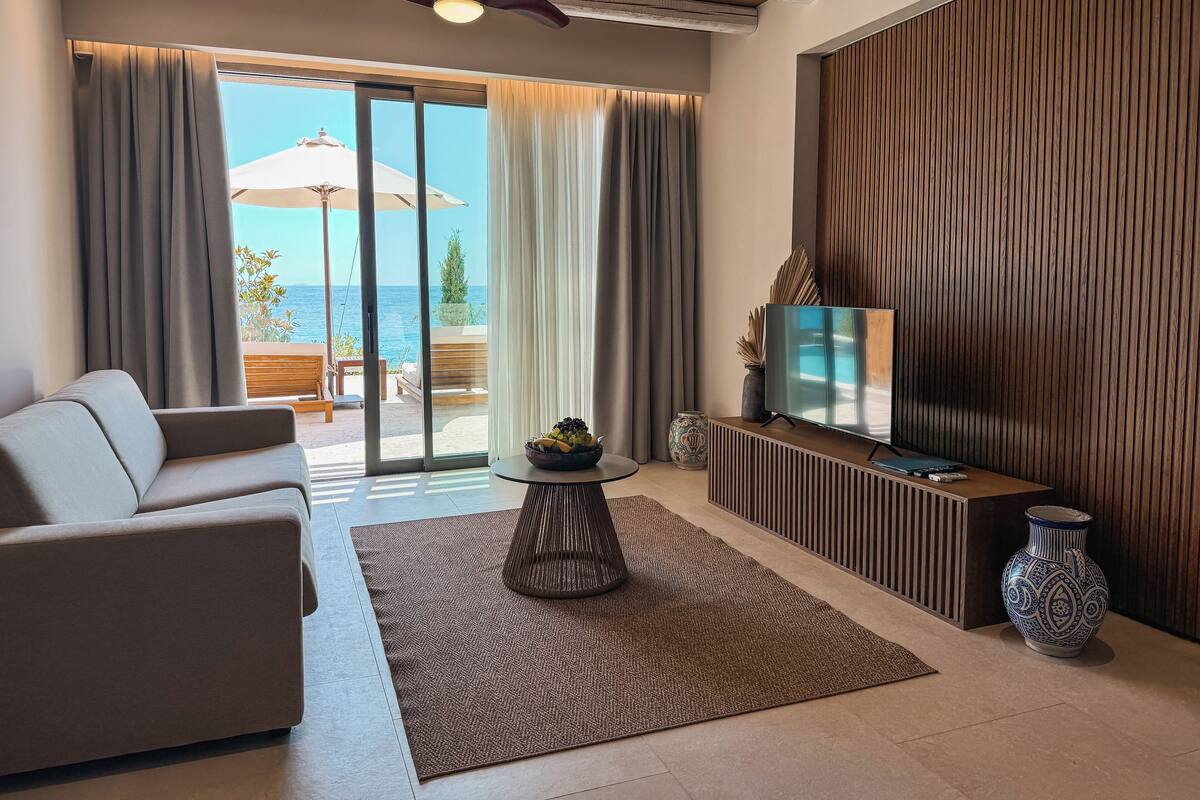 Een moderne woonkamer met een bank, een televisie en uitzicht op de zee door grote ramen.