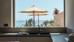 Eine Küche mit einem großen Fenster zum Meer, einem beigen Sonnenschirm und einer Holzwand mit Pflanzen.