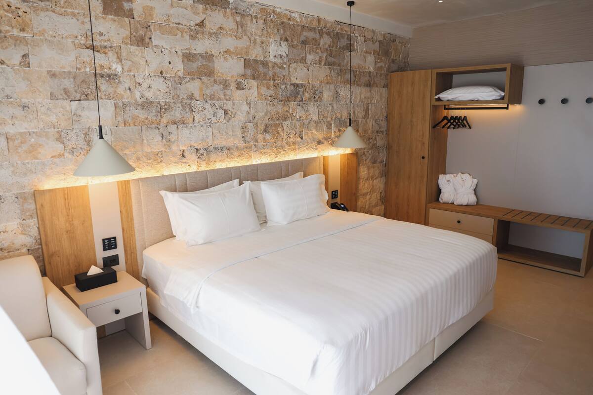 Een moderne slaapkamer met een stenen muur, een groot bed met witte lakens, twee bedlampjes, een houten nachtkastje, een houten bankje en een ingebouwde plank met een badjas en handdoek.