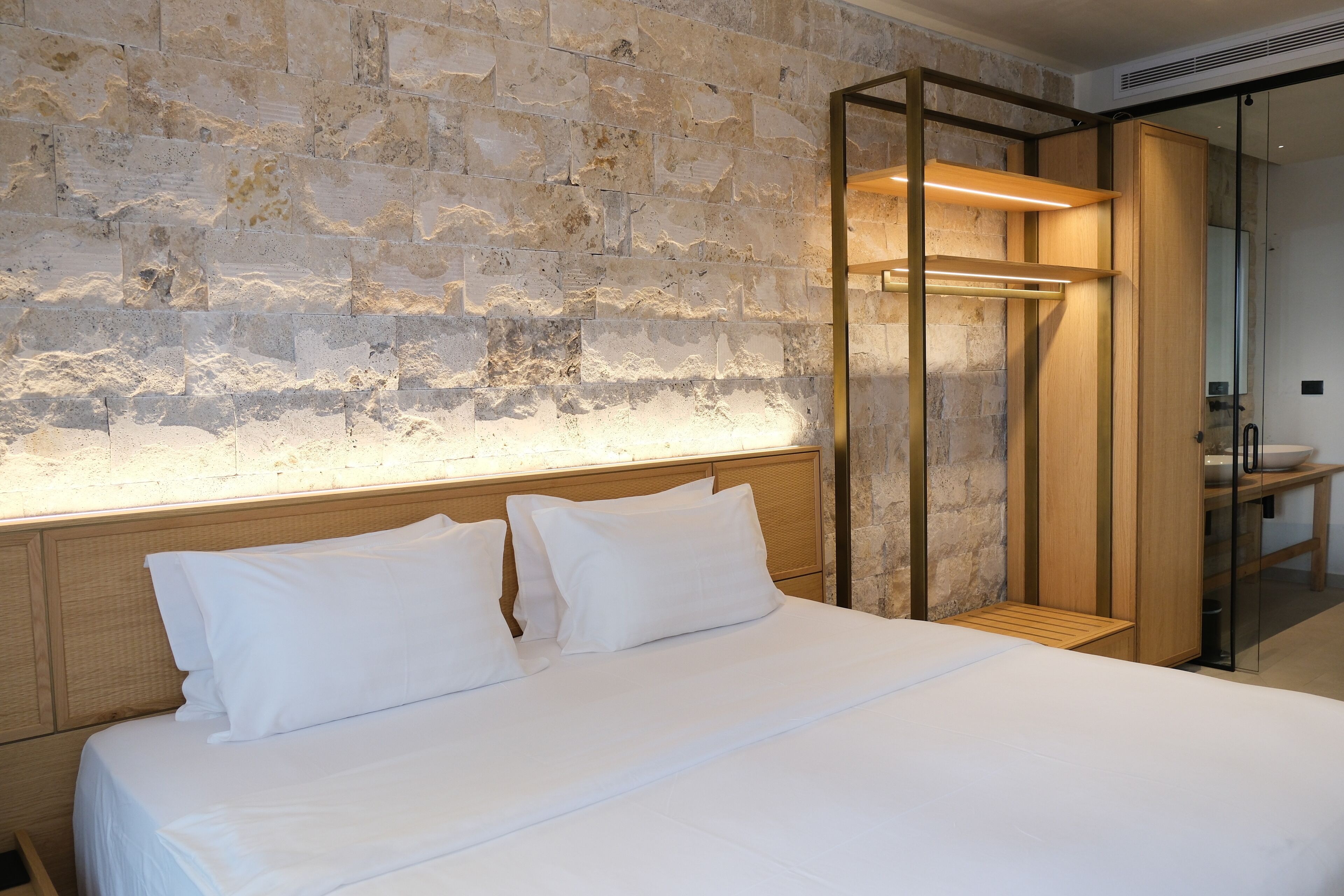 A modern hotel room with a stone wall, a wooden headboard, and a glass shower enclosure.