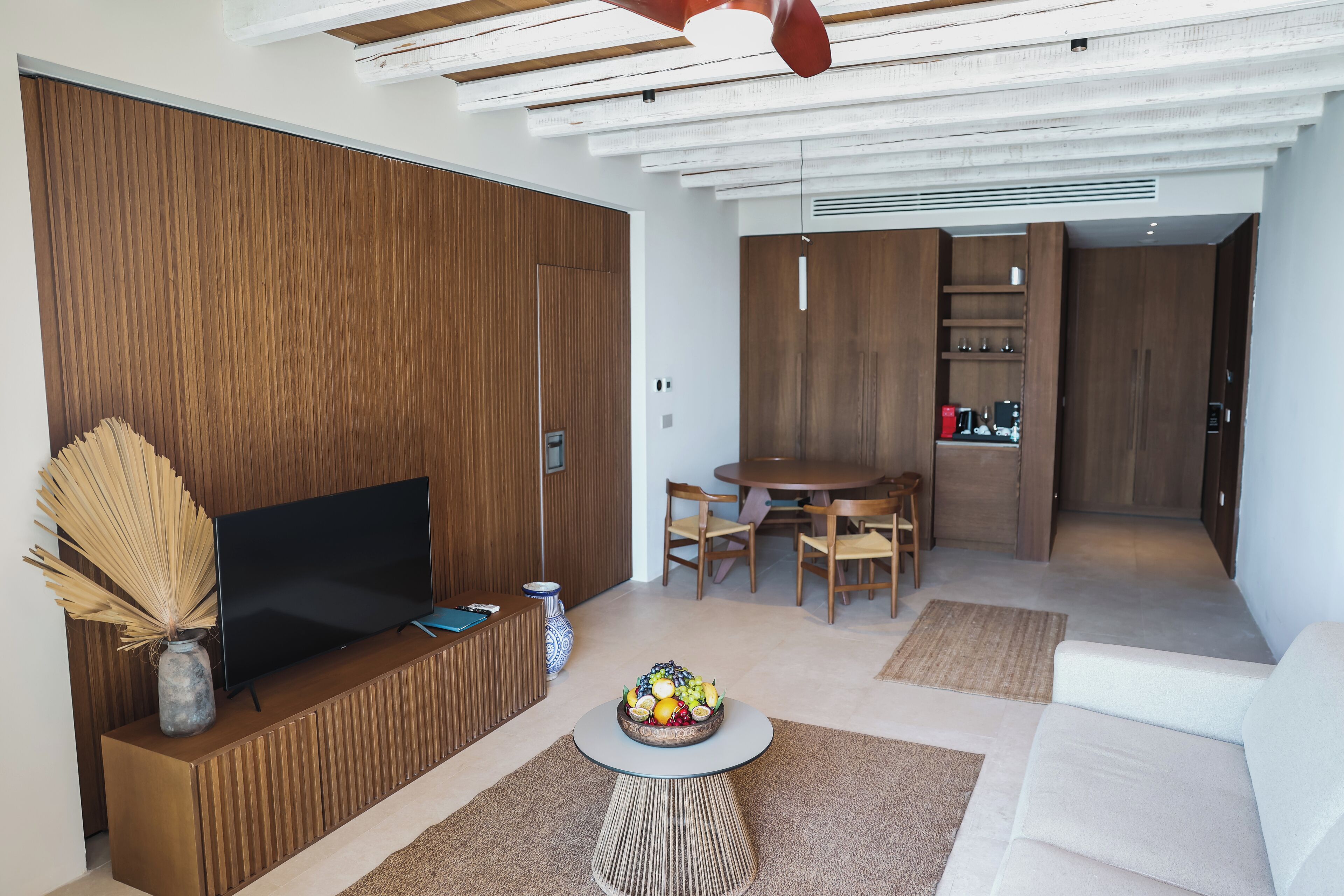 Een moderne woonkamer met een flatscreen televisie, houten lambrisering, een eethoek met een ronde tafel en stoelen, en een witte bank.