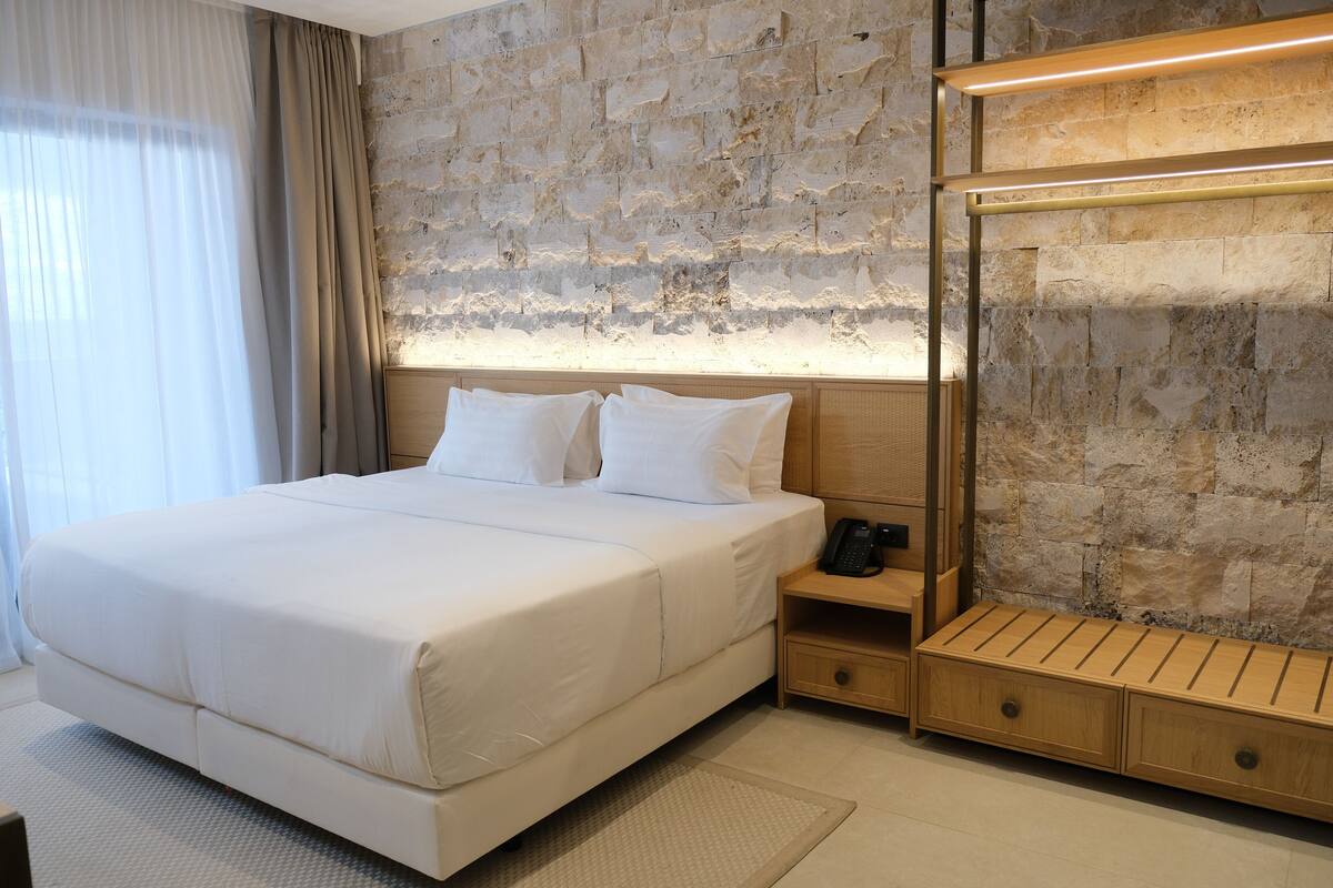 Een moderne hotelkamer met een stenen muur, een groot bed met witte lakens, een nachtkastje met een telefoon en een houten bankje.