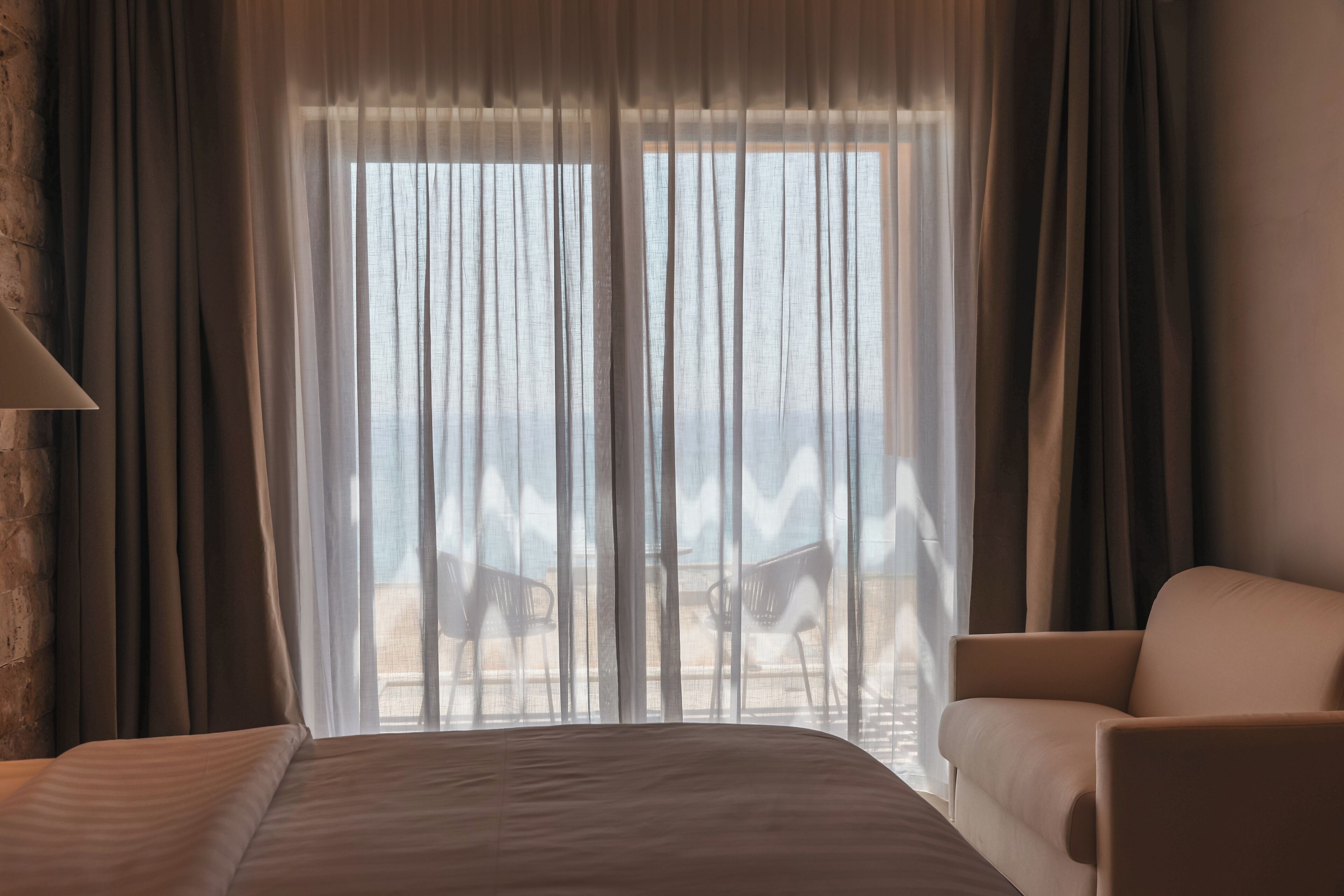 Habitación de hotel con una cama grande, una silla y vistas a la playa a través de cortinas translúcidas.