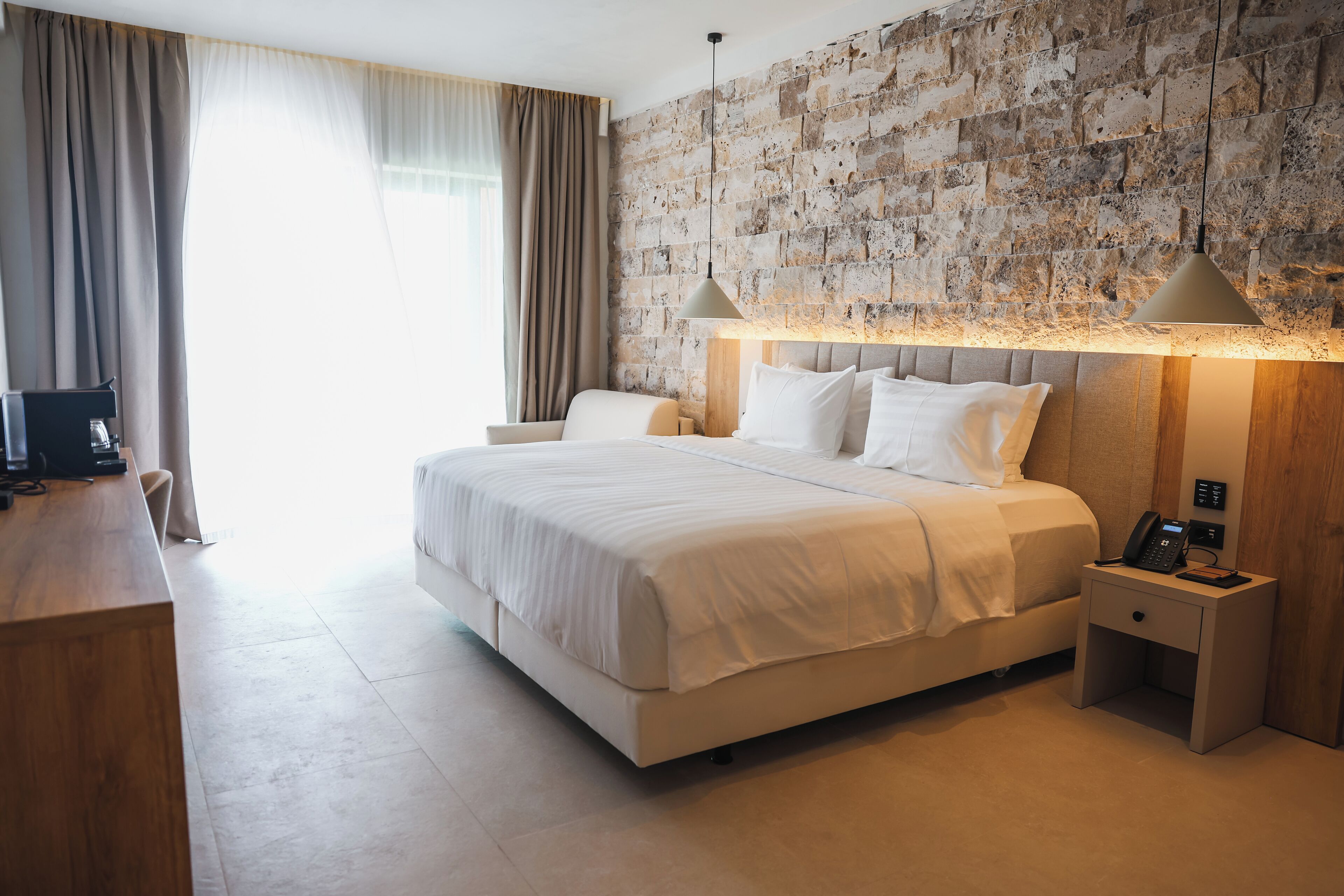 A modern hotel room with a large bed, bedside lamps, a wooden desk, and a stone-textured wall.