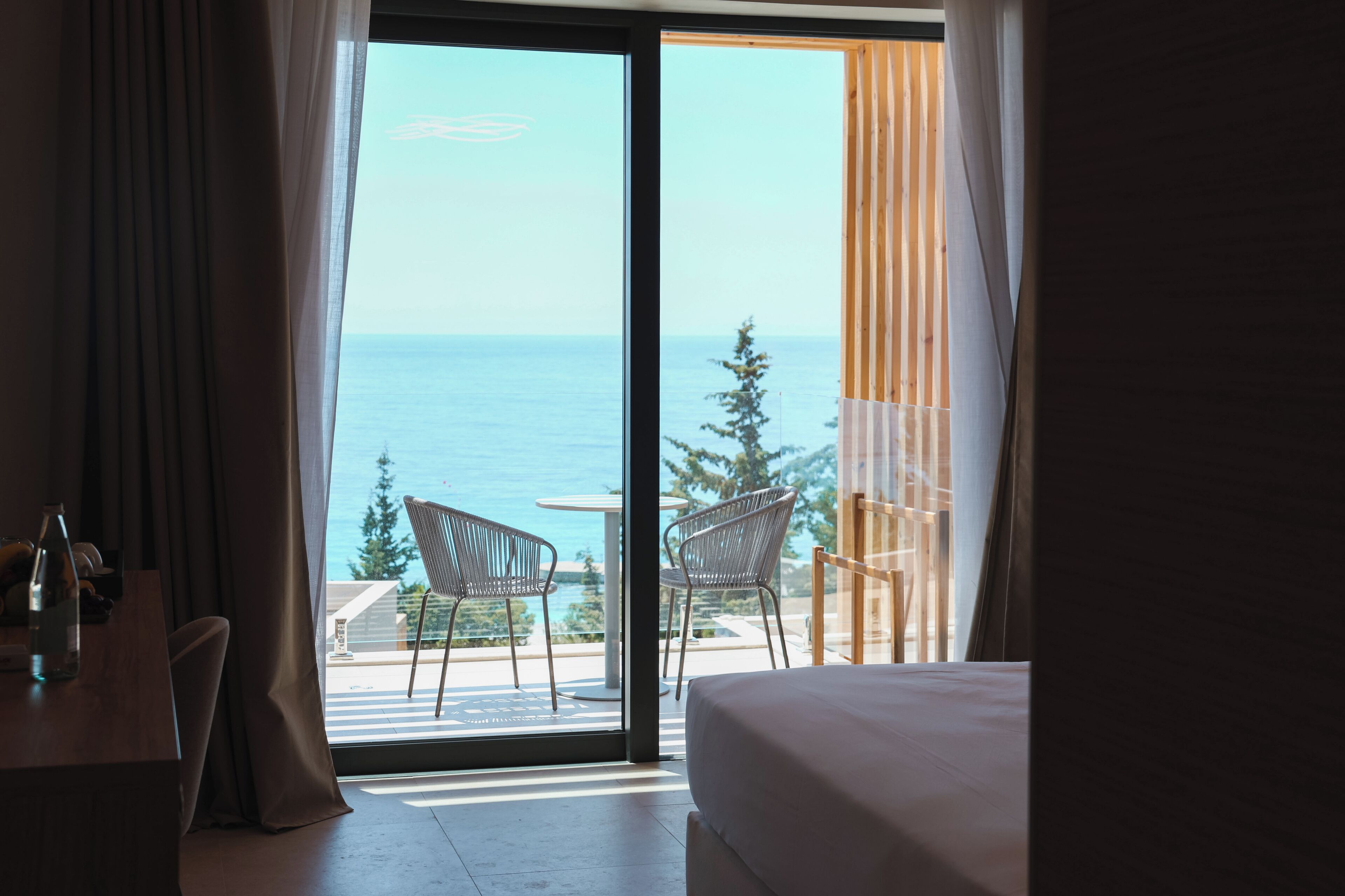Habitación de hotel con un gran ventanal corredero que da a un balcón con vistas al mar y a árboles.