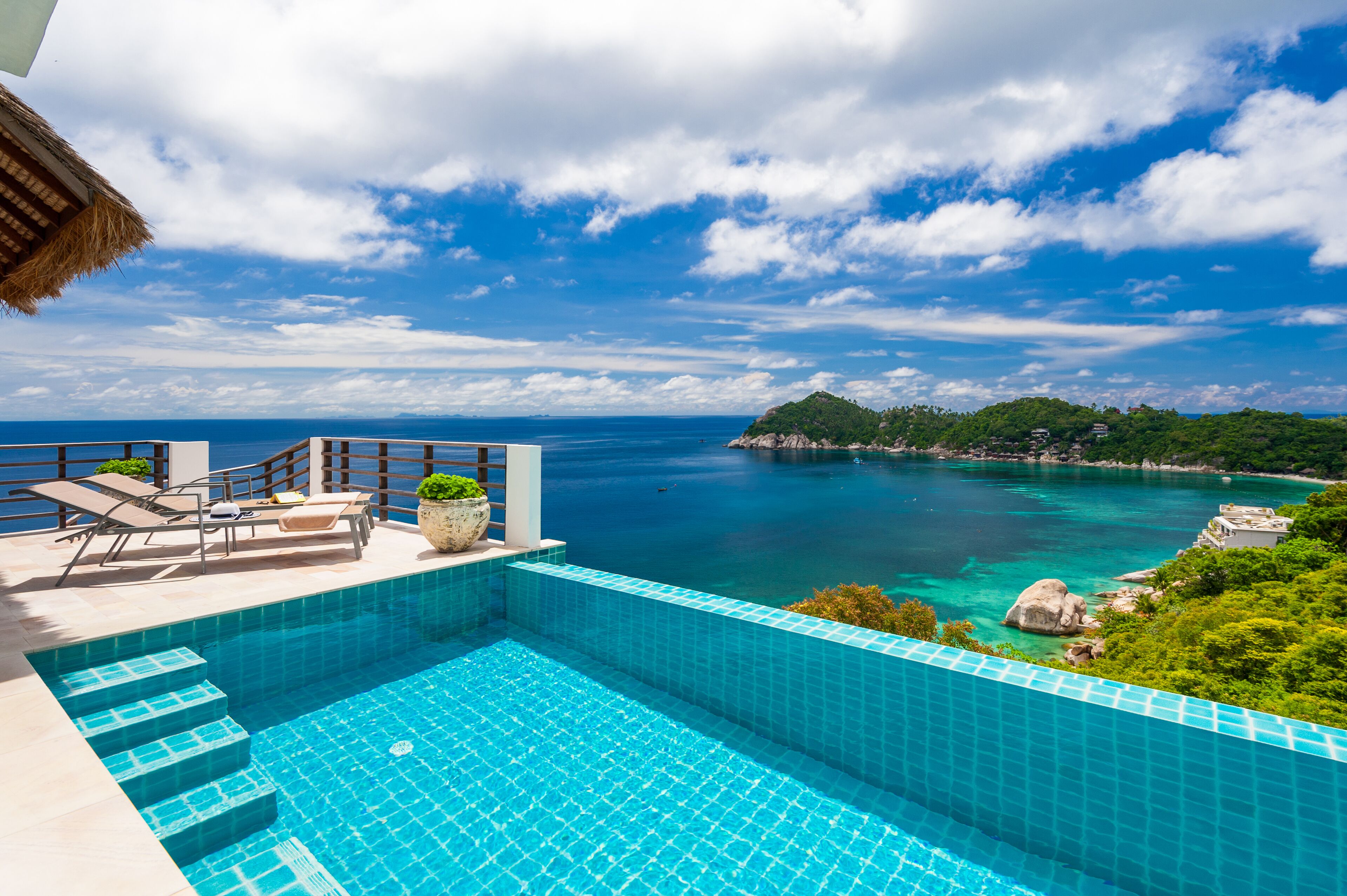 A swimming pool with steps leading into it, lounge chairs, and a view of the sea and greenery.