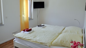 A hotel room with a bed, a flat-screen TV mounted on the wall, a window with yellow curtains, and a small table with a remote control. - Modernes Apartmenthaus (Falkensee)