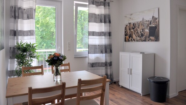 A dining area with a wooden table and chairs, a vase with flowers, and a black trash bin. - Modernes Apartmenthaus (Falkensee)