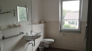 A bathroom with a toilet, sink, and mirror. There is a window providing natural light and a view of a house outside. - Modernes Apartmenthaus (Falkensee)