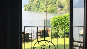 A view from a room with a door leading to a balcony overlooking a lake, with a grassy area and a metal fence. - Ossuccio Villa Darsena private lakeside villa with pool by Italian Apartments. (Ossuccio)