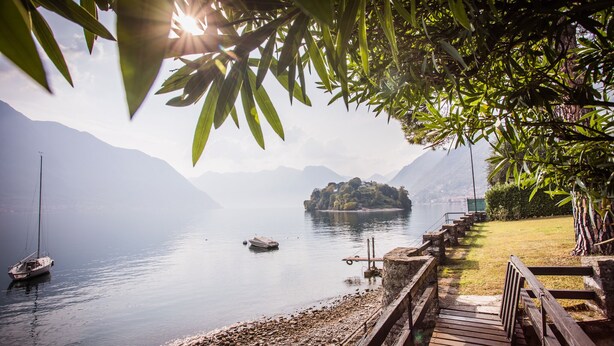 A wooden deck leading to a lake with boats, a distant island, and mountains in the background. - Ossuccio Villa Darsena private lakeside villa with pool by Italian Apartments. (Ossuccio)