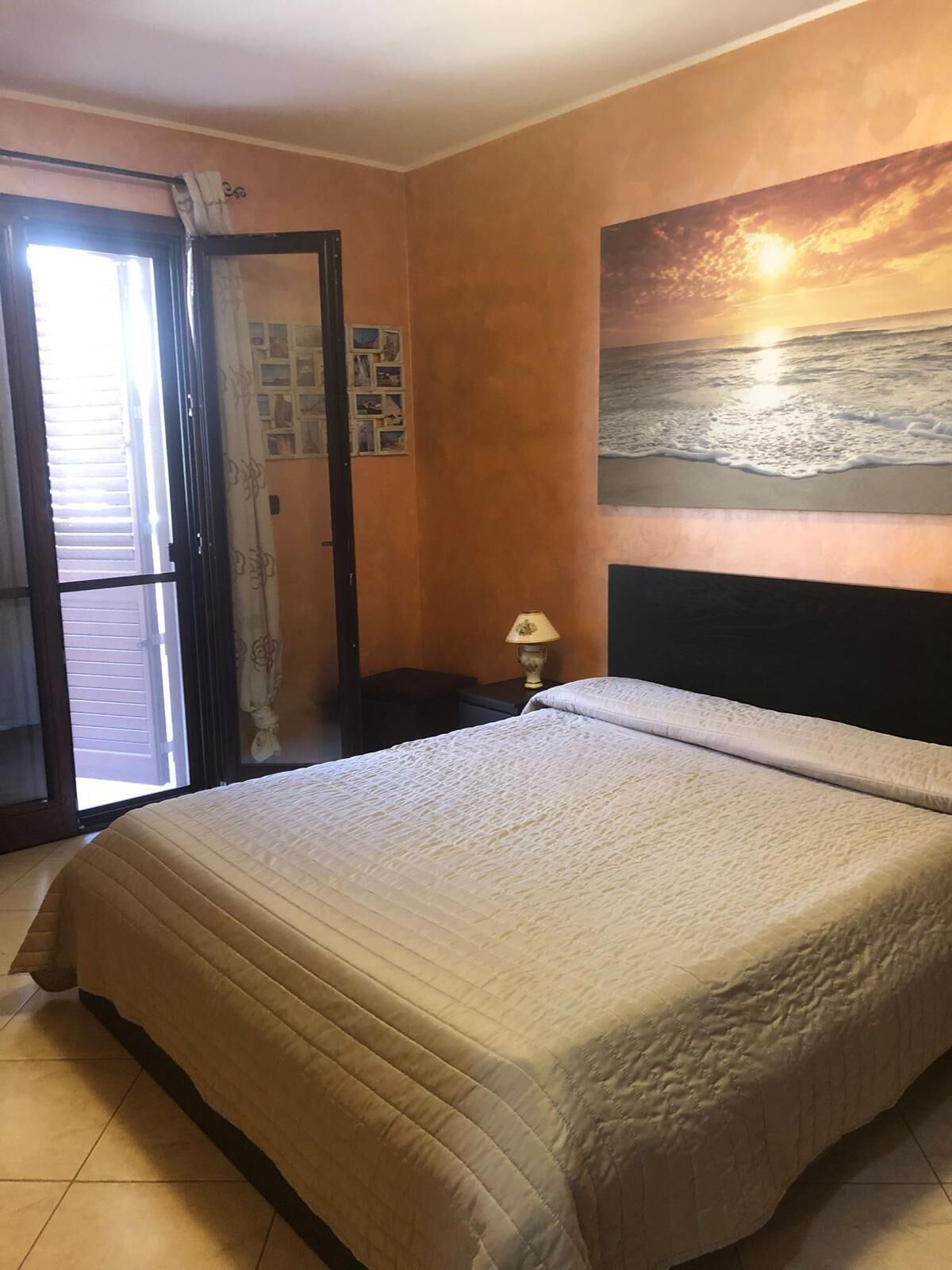 Un dormitorio con una cama, una mesita de noche, una lámpara, un cuadro de un atardecer y una puerta que da a un balcón.