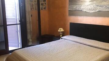 Un dormitorio con una cama, una mesita de noche, una lámpara, un cuadro de un atardecer y una puerta que da a un balcón.