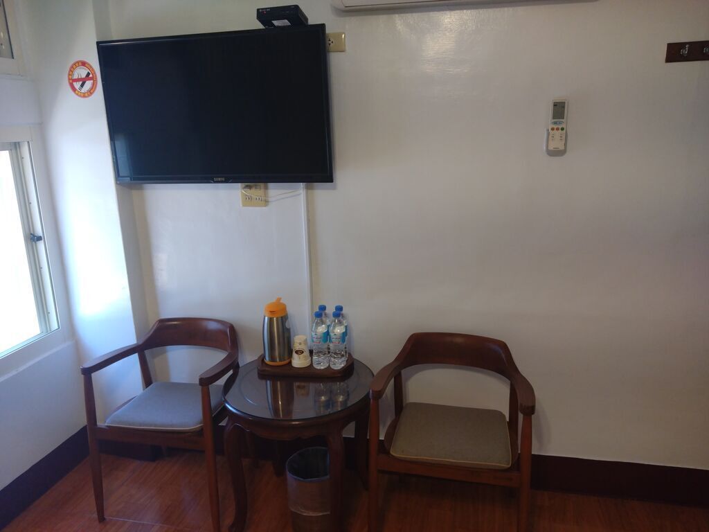A room with a television mounted on the wall, a small table with chairs, and a trash bin.