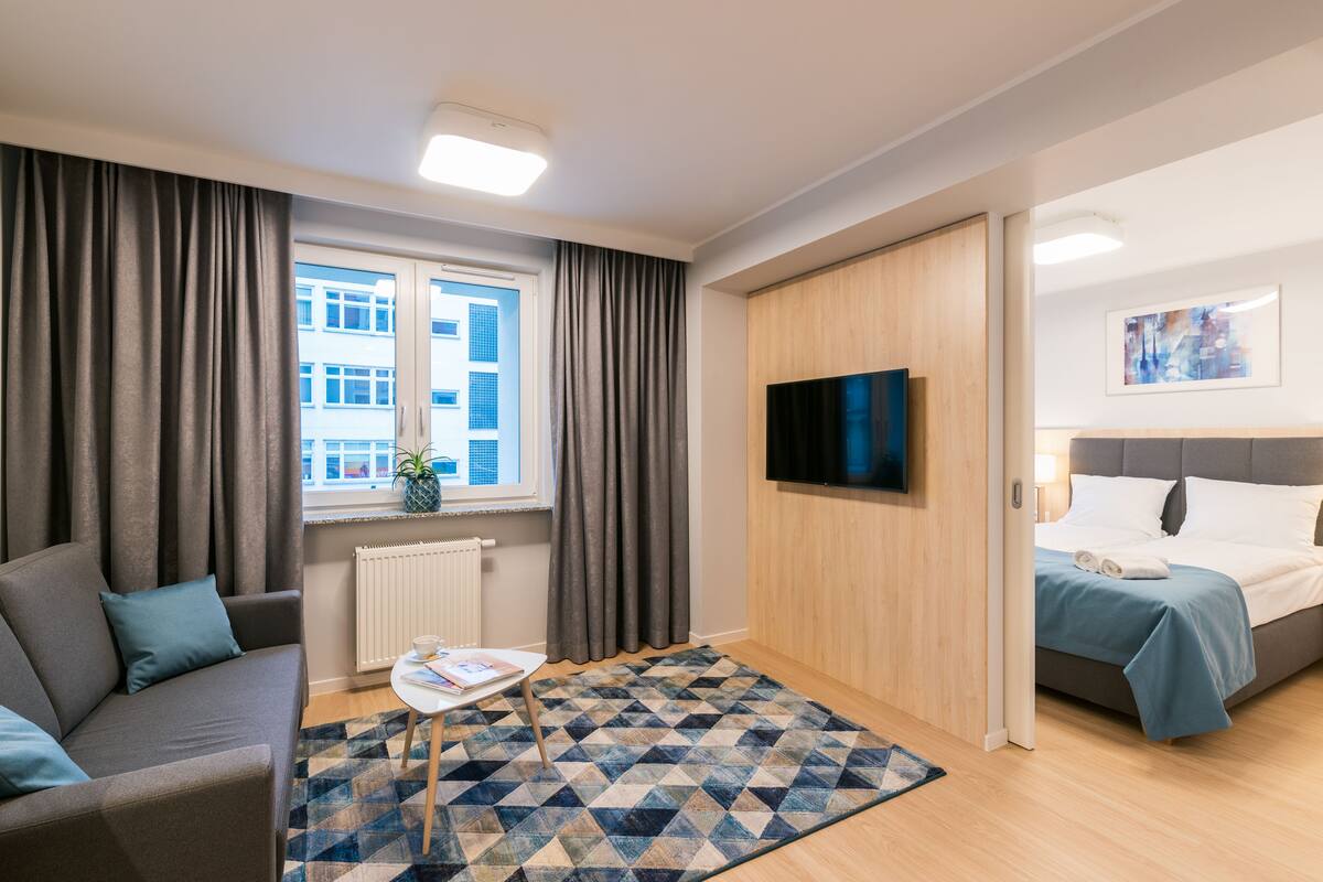 Une chambre d’hôtel moderne avec un canapé, une petite table sur laquelle repose un livre, un tapis à motifs, un lit recouvert d’une courtepointe bleue, un téléviseur fixé au mur et une fenêtre avec des rideaux.