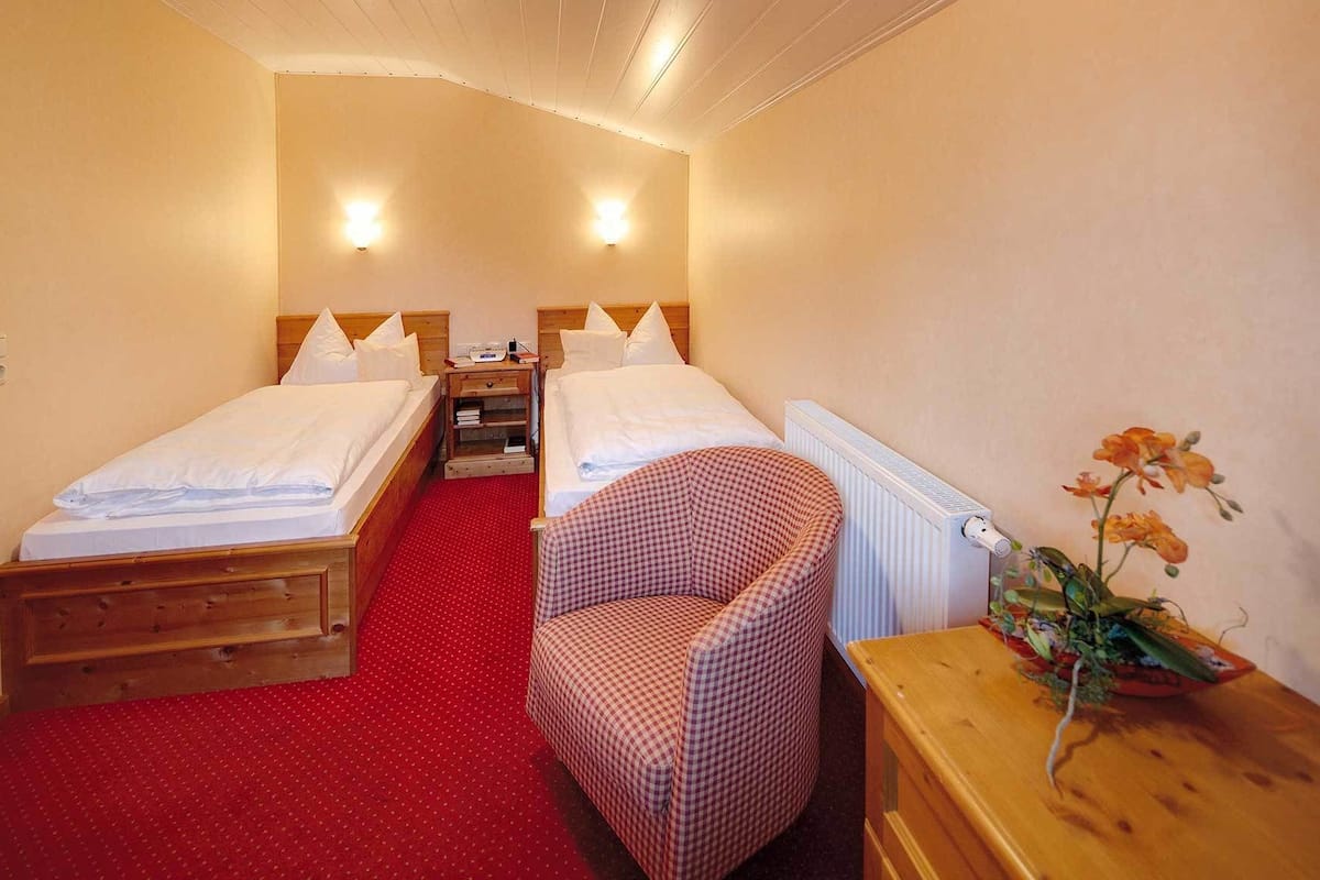 A hotel room with two single beds, a red carpet, a checkered chair, a wooden nightstand, and a vase with flowers.