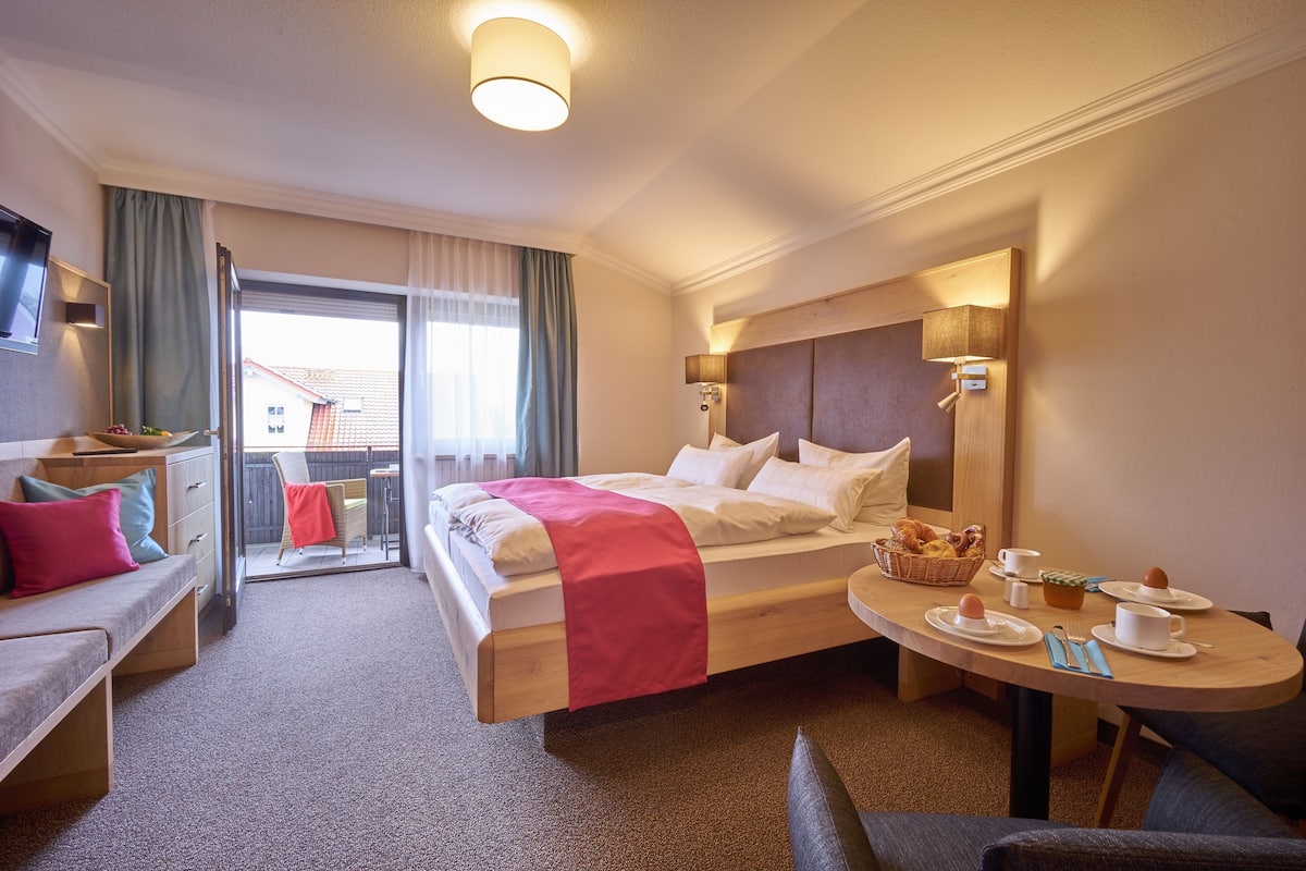 A hotel room with a bed, a desk, a dining table set for breakfast, and a balcony with a view.