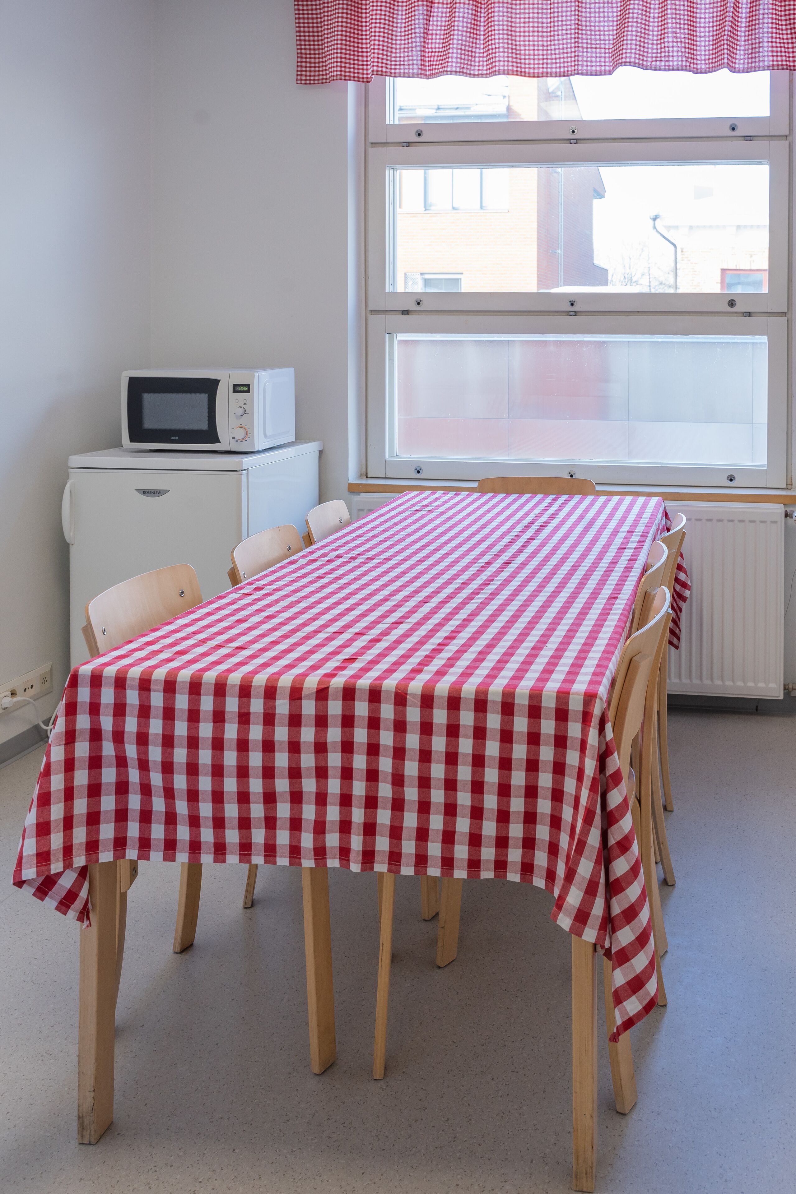 Una stanza con un tavolo a scacchi, sedie, un termosifone bianco e un forno a microonde su un piccolo mobile bianco.