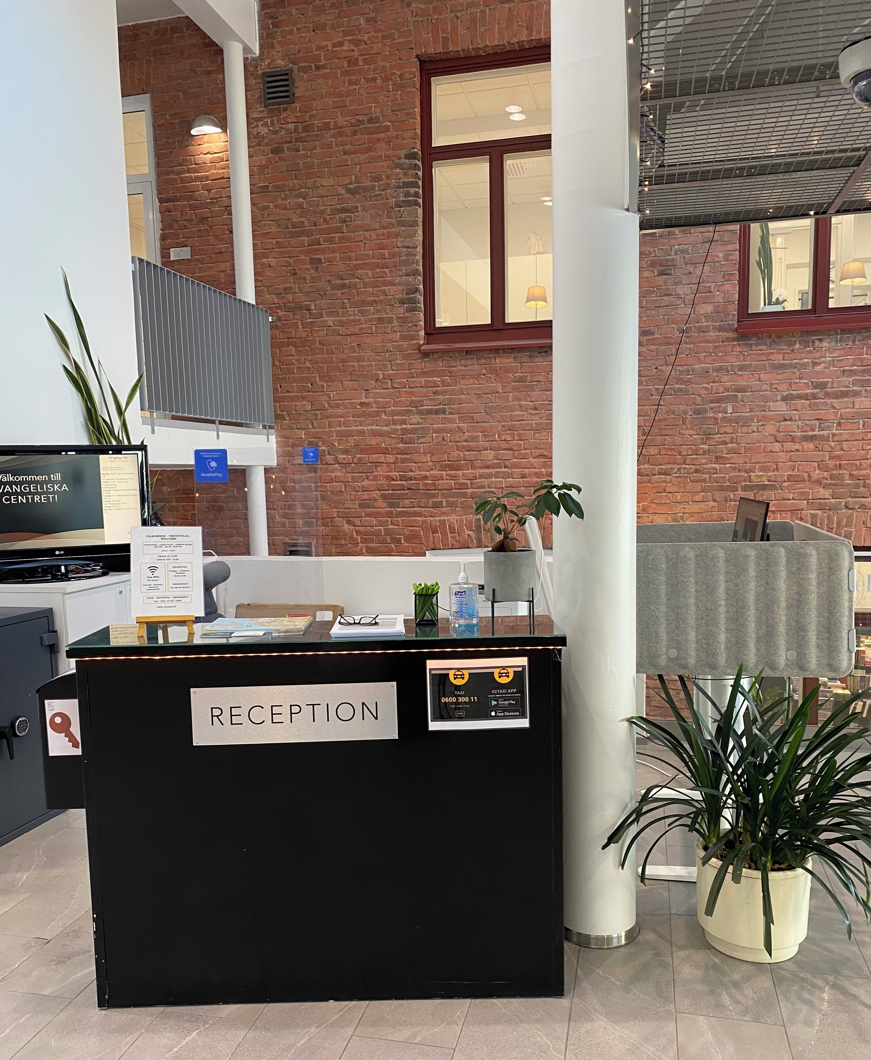 A reception desk with a sign labeled "RECEPTION," a computer monitor, a plant, and a window with a view of the outside.