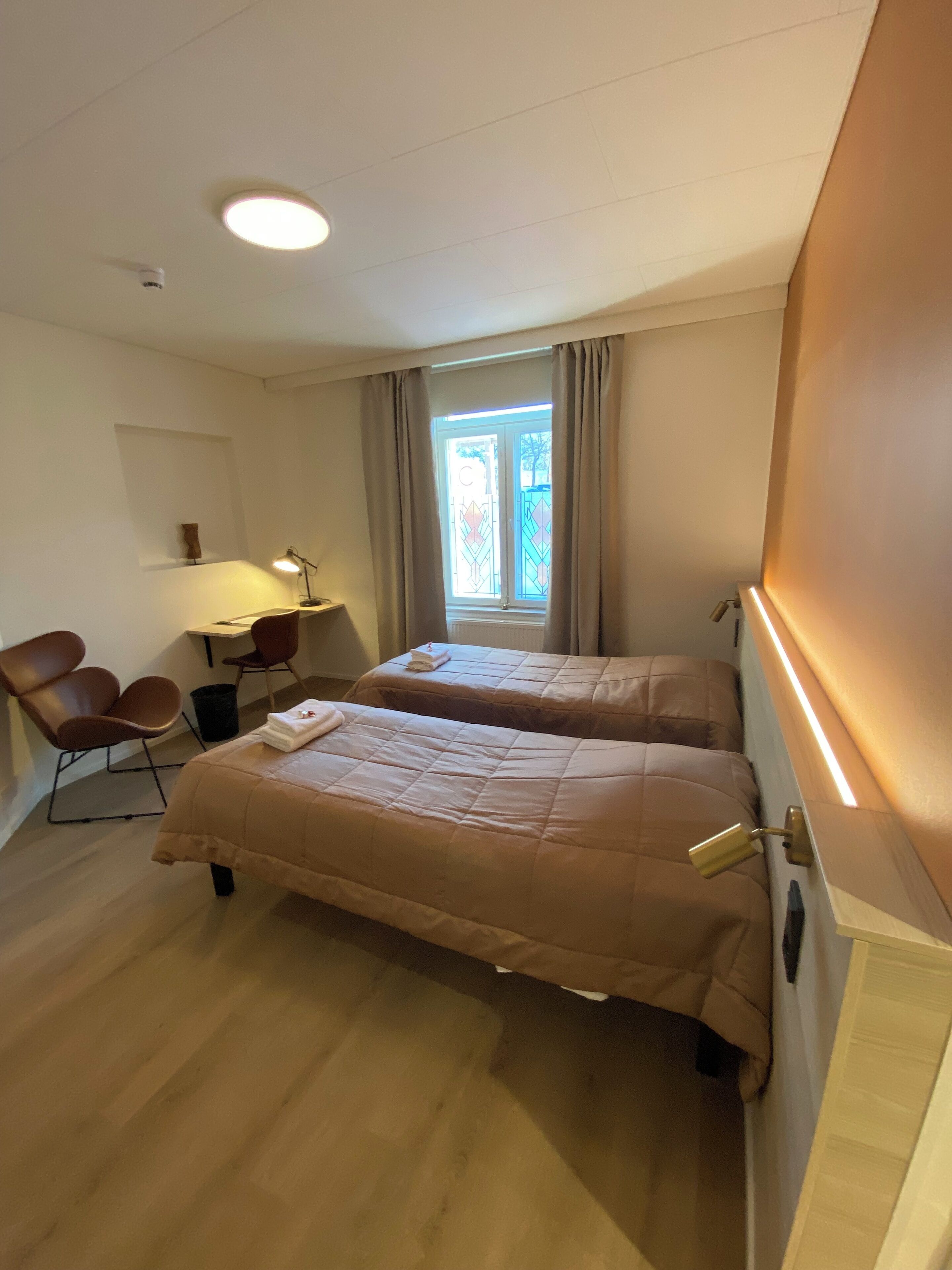Habitación de hotel con dos camas, un escritorio con lámpara, una silla y una ventana con cortinas.