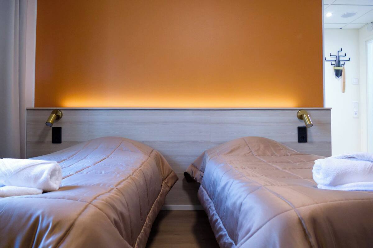 Quarto de hotel com duas camas, cabeceira de madeira e luminária fixada na parede.