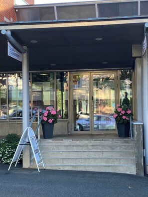 Une entrée d’immeuble avec des portes vitrées, un porche couvert et deux plantes en pot ornées de fleurs roses.