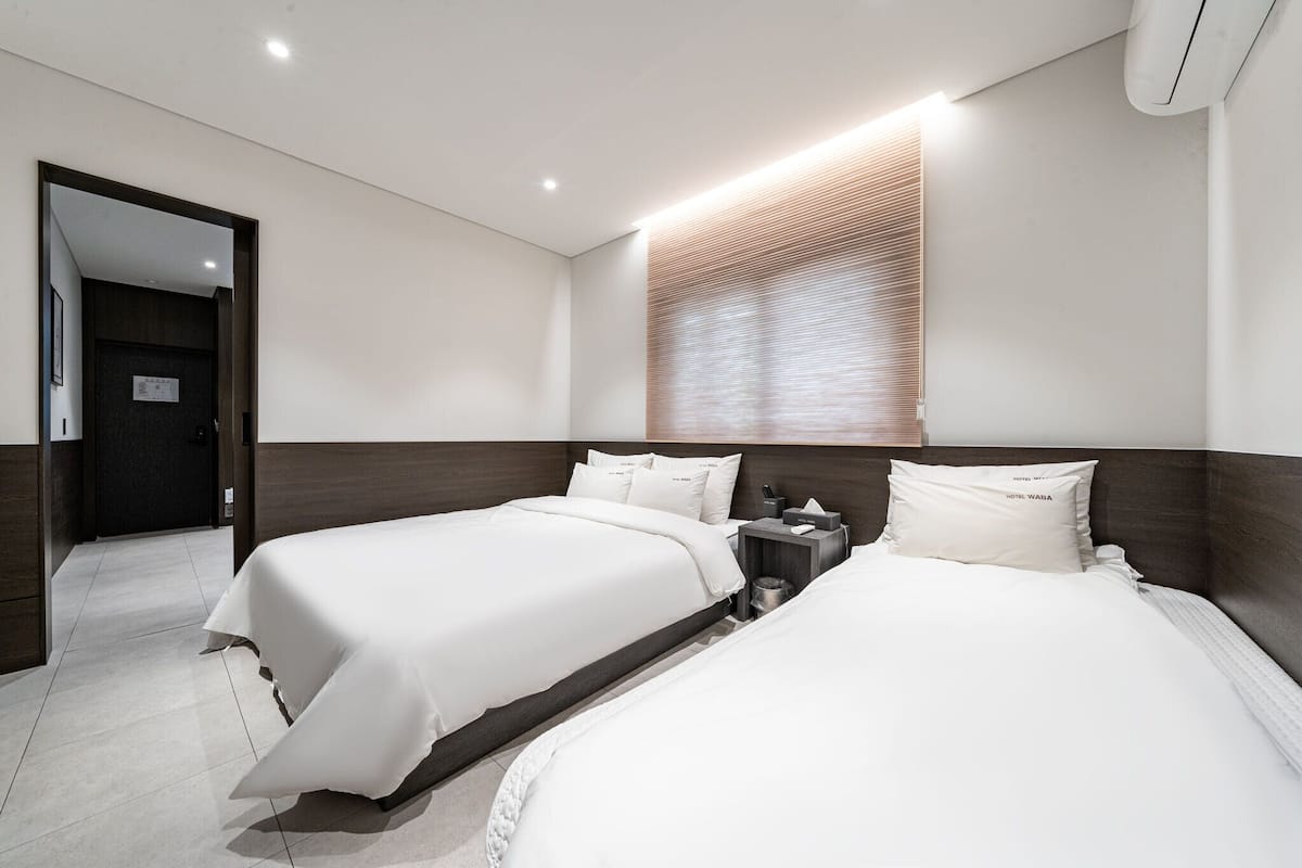 A modern hotel room with two beds, a wooden headboard, a bedside table with a lamp, and a large window with blinds.