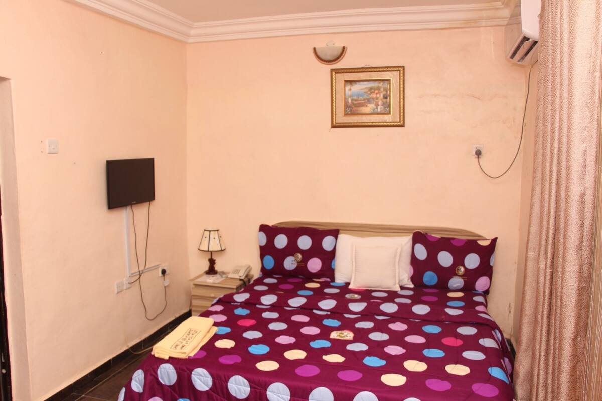 A bedroom with a polka dot bedspread, a flat-screen TV mounted on the wall, a bedside lamp, and a framed picture above the bed.