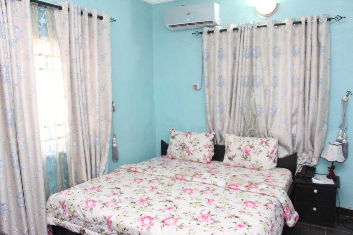 A bedroom with a floral bedspread, a bedside table with a lamp, and curtains on the windows.
