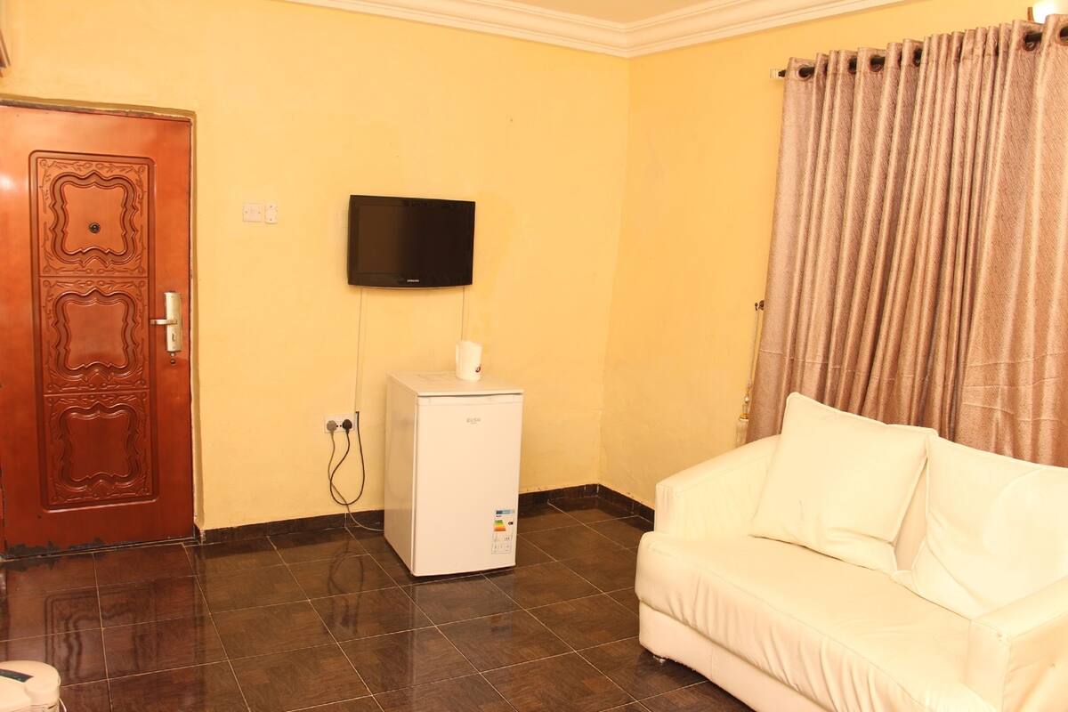 A room with a brown door, a white refrigerator, a flat-screen TV mounted on the wall, and a beige sofa.